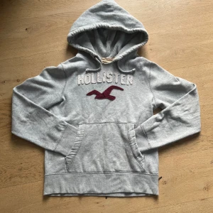 Grå vintage hoodie från hollister - Snygg vintage grå hoodie från Hollister med stor broderad logga och fågel i vinrött på bröstet. Tröjan har huva med snörning, känguruficka och långa ärmar. Perfekt för chill dagar och enkel att matcha med jeans eller joggers.