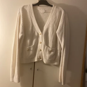 Vit kofta från H&M - Säljer en vit ribbstickad kofta från H&M i storlek S. Koftan har guldiga knappar framtill och på fickorna, v-ringad hals och två små fickor. Perfekt för lager-på-lager och enkel att styla till olika outfits.