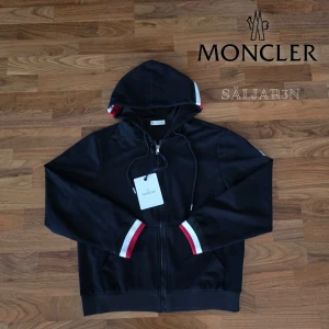 Moncler Hoodie - Exklusiv Moncler hoodie i svart, storlek S. Nyskick med taggar! Elegant design med hel dragkedja och klassiska Moncler-detaljer i rött, vitt och blått på muddar och huva. Ikoniskt logomärke på ärmen för en exklusiv touch. Huva med dragsko och fickor för en modern, funktionell stil. Tillverkad i mjukt och slitstarkt tyg som ger både komfort och premiumkänsla. En tidlös lyxig hoodie som lyfter varje outfit. 📦 Snabb leverans – skickas samma dag!