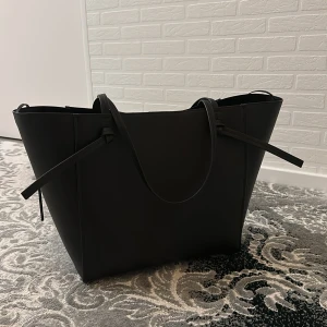 Svart stor axelväska i skinn - Snygg svart axelväska med minimalistisk design och stilrena linjer. Väskan är tillverkad i slätt skinn och har långa handtag som gör den enkel att bära över axeln. Perfekt för dig som gillar en clean look och vill ha plats för allt du behöver.