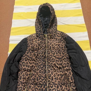 Leopardmönstrad pufferjacka  - Säljer en fet pufferjacka från Rainbow med leopardmönster på kroppen och svarta ärmar. Jackan har huva, guldfärgade dragkedjor och två fickor framtill. Perfekt för dig som vill sticka ut i höst och vinter. Materialet är syntet och jackan är riktigt varm.