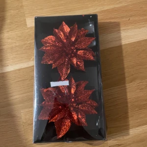 Röda glittriga blomdekorationer - Två stora röda blommor täckta av glitter, formade som julstjärnor och förpackade i en svart ask. Perfekta som dekoration till jul eller fest. Materialet är konstgjort och blommorna har en tydlig glanseffekt.