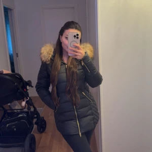 Svart pufferjacka  - Snygg svart pufferjacka från Rockandblue med guldig dragkedja och detaljer. Jackan har en stor huva med fluffig i  beige/brun ton och quiltad design. Perfekt för kalla dagar och ger en cool siluett.