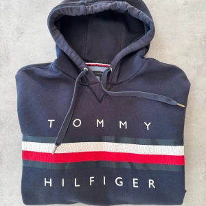 Marinblå hoodie Tommy Hilfiger M - Snygg marinblå hoodie från Tommy Hilfiger i storlek M. Tröjan har huva med snörning, klassisk logga och breda ränder i vitt, rött och grått över bröstet. Mjuk bomullskvalitet och känguruficka framtill. Perfekt för chill dagar och streetstyle.