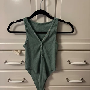 Ribbad grön ärmlös bodysuit - Snygg ribbad bodysuit i dämpad grön färg med v-ringning och tryckknappar framtill. Ärmlös modell som sitter tight och har en cool, enkel look. Perfekt att styla med jeans eller kjol för en trendig outfit.