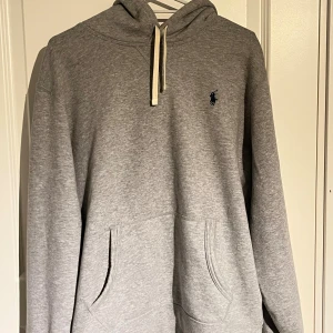 Grå hoodie från Ralph Lauren - Säljer en stilren grå hoodie från Ralph Lauren med klassisk huva och vit snörning. Tröjan har en broderad logga i mörkblått på bröstet. Jag skulle säga att den passar mer för dem som har storlek S men den funkar också för dem som har M.