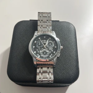 Stilren silverfärgad klocka med länk - Säljer en snygg klocka i silverfärg med robust länkarmband och svart urtavla med flera detaljerade visare och kugghjul. Klockan har en klassisk rund form och metallarmband som ger en modern och cool look.