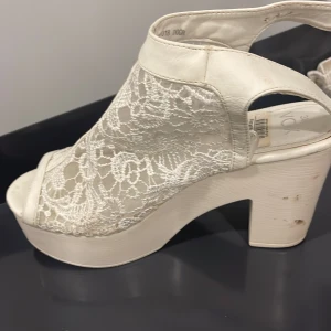 Vita spetsiga sandaler med klack från VOX - Säljer ett par vita sandaler från VOX i storlek 38. Skorna har en chunky klack och plattform, öppen tå och är täckta med snygg spetsdetalj. De har ett spänne vid hälen och är perfekta för dig som gillar en romantisk och trendig look.