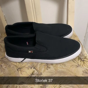 Svarta slip-on sneakers Tommy Hilfiger - Svarta slip-on sneakers från Tommy Hilfiger med vit sula och diskret logga på sidan. Skorna har en enkel och stilren design i textilmaterial, perfekt för en avslappnad och snygg look.