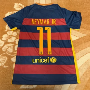 FC Barcelona Neymar Jr matchtröja - Säljer en officiell FC Barcelona matchtröja från Nike med Neymar Jr och nummer 11 på ryggen. Tröjan har breda blå och röda ränder, gula detaljer och klubbmärke på bröstet. Tillverkad i lätt och ventilerande polyester med Dri-FIT-teknologi.