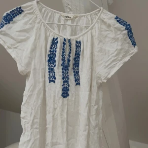 Blus med broderi - Superfin vit blus med blått broderat blommönster på axlar och framtill💙Blusen har korta ärmar, rund halsringning och en liten knapp framtill. Storlek 170 men passar mig som vanligtvis bär xs-s perfekt. Den har en ytterst liten fläck som knappt syns (sista bilden), men den tvättar jag bort vid försäljning. Nästan helt oanvänd så är i jättebra skick annars🌸
