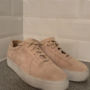 Beige sneakers från Axel Arigato -  Skorna ser smutsigare ut än va de är men de är ett par Snygga beige sneakers från Axel Arigato i mjuk mocka med vit platt sula. Klassisk låg modell med rund tå och snörning. Perfekt för dig som gillar stilrena och minimalistiska skor med premiumkänsla.