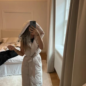 Lång beige dunväst med huva och bälte - Super fin. Passar XS - M. Justerbart med band hur tajt du vill ha den. Har även knappar. 