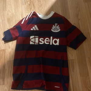Newcastle United borta ställ 24/25  - Snygg Newcastle United fotbollströja från Adidas med breda marinblå och vinröda ränder, vit krage och tryckt sponsorlogga på bröstet. Kortärmad modell i lätt och ventilerande Aeroready-material, perfekt för match eller träning.