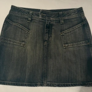 Svart jeanskjol från Lindex - Cool svart jeanskjol från Lindex med slitna detaljer och stora fickor både fram och bak. Kjolen har bälteshällor, dragkedja och knapp framtill. Klassisk rak modell i denim som är enkel att styla med allt.