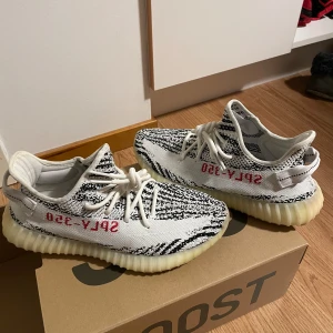 Adidas Yeezy Boost 350 Zebra - Säljer ett par Adidas Yeezy Boost 350 V2 'Zebra' med ikoniskt svartvitt randigt mönster, röd SPLY-350 text på sidan och vit, ribbad sula. Skorna har snörning och är tillverkade i syntetmaterial med bekväm boost-sula. Kommer med originalkartong.