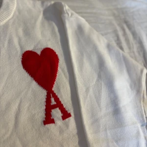 Vit stickad tröja från Ami Paris - Säljer en vit stickad tröja från Ami Paris med ett stort rött hjärta och bokstaven A broderat på bröstet. Tröjan har rund hals, ribbade muddar och är långärmad. Perfekt för dig som gillar stilrena detaljer och vill sticka ut med en ikonisk design.