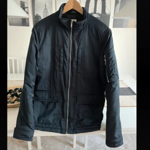 Svart bomberjacka Jack & Jones M - Svart bomberjacka från Jack & Jones Premium, storlek M. Jackan har ståkrage, dragkedja framtill, fickor med lock och en extra ficka med dragkedja på ärmen. Materialet är polyester och jackan har en stilren, klassisk look.