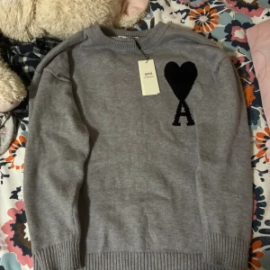 Grå stickad tröja från Ami Paris - Snygg grå stickad tröja från Ami Paris med svart hjärta och A-logga på bröstet. Tröjan har rund hals, ribbade muddar och är tillverkad i ekologisk bomull. Perfekt för dig som gillar stilrena och trendiga plagg. Passar bra för S/M