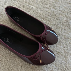 Vinröda ballerinaskor med rosett - Snygga vinröda ballerinaskor från Clou med glansig finish och rund tå. Dekorativa rosetter framtill med guldfärgade detaljer ger en extra touch. Perfekta att matcha med jeans eller klänning för en chic look.