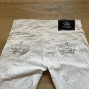 Victoria Beckham jeans - Snygga vintage Victoria Beckham jeans från Rock & Republic med raka ben och broderade kronor på bakfickorna. Byxorna har silverfärgade nitar och klassisk femficksdesign. Perfekta för dig som vill sticka ut med en clean och lyxig look. Low waist och bootcut. Storlek w29. 