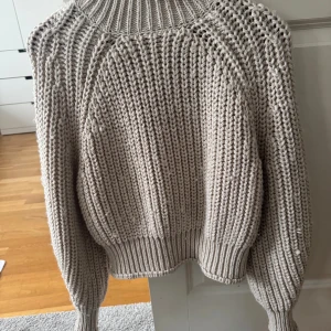 Beige stickad tröja från H&M - Säljer en chunky beige stickad tröja från H&M i storlek S. Tröjan har bred ribbad struktur, hög krage och långa ärmar med extra breda muddar. Perfekt för dig som gillar en cozy och trendig look med lite oversized känsla.