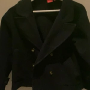 Svart kappa från RYVLS - Stilren svart skepparkavaj från RYVLS med bred krage och dubbla rader av guldknappar framtill. Jackan har en klassisk, rak passform och är perfekt för dig som gillar en tidlös look. Snygg att matcha med jeans eller chinos.