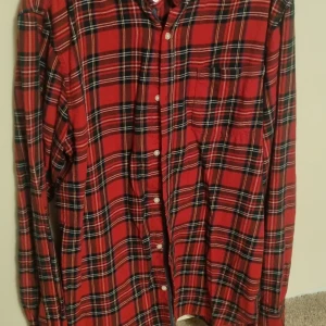 Röd rutig (tartan) skjorta från Next - Snygg röd skjorta från Next med klassiskt rutigt mönster i svart, vitt och gult. Skjortan har långa ärmar, knappar framtill och en bröstficka. Perfekt för dig som gillar en chill och tidlös stil. Materialet är mjukt och känns som bomull. Storlek 176/16 år