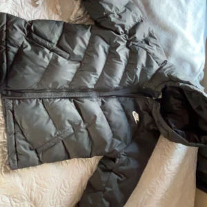 Svart pufferjacka från Nike med huva - Svart pufferjacka från Nike med huva och dragkedja framtill. Jackan har quiltad design med breda paneler och Nike-logga på bröstet. Perfekt för kalla dagar och har en clean, sportig look. Materialet är syntet och jackan är långärmad.