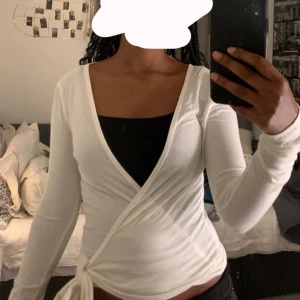 Vit omlottopp med knyt i sidan - Säljer en vit omlottopp med lång ärm och snygg v-ringning. Toppen har knyt i sidan och sitter figurnära. Perfekt att styla med jeans eller kjol.Använt 1 gång