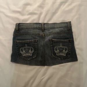 Jeanskjol med broderade kronor - Superkort vintage jeanskjol i klassisk blå denim från fem-cace. Två bakfickor med snygga broderade kronor i vitt ger kjolen en unik touch. Gylf med dragkedja och knapp, samt bälteshällor. Perfekt för dig som gillar streetstyle och detaljer. Står storlek 38 men skulle säga 34/36