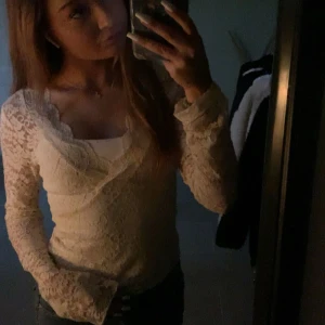 Vit spetsblus med omlott och lång ärm - Säljer en vit blus i spets med omlott-design och långärmade ärmar. Blusen har en v-ringad hals och är figurnära med transparent spets över hela plagget. Perfekt för dig som gillar romantisk och feminin stil.