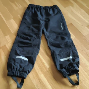 Svarta täckbyxor Kaxs Proxtec stl 110 - Svarta täckbyxor från Kaxs Proxtec i storlek 110. Byxorna har resår i midjan och benslut, reflexdetaljer på benen och justerbara fothällor. Perfekta för utomhuslek och håller dig torr och varm under kalla dagar.