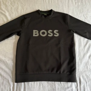 Svart sweatshirt från BOSS - Svart sweatshirt från BOSS med stor grå logotyp på bröstet. Tröjan har rund hals och långa ärmar, tillverkad i mjukt material som känns skönt mot huden. Perfekt för dig som gillar stilrena och enkla plagg med tydlig branding.