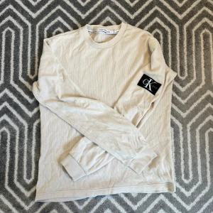 Beige långärmad tröja Calvin Klein - Snygg beige långärmad tröja från Calvin Klein Jeans med ribbade muddar och ett stort svart CK-märke på ärmen. Tröjan är storlek XS men passar S. Tveka inte på att skriva om du undrar nåt!✨