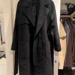Svart kappa från 2NDDAY, stl 40 - Stilren svart kappa från 2NDDAY med bred krage, knappar framtill och tillhörande bälte i midjan. Lång modell med raka ärmar och två fickor framtill. Perfekt för dig som gillar en klassisk och clean look.