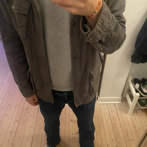  Woolrich jacka  - Parkajacka från Woolrich i grått med flera stora fickor framtill och axeldetaljer. Jackan har lång ärm, knappar och dragkedja samt patch med logga på ärmen. Materialet är slitstark bomull, perfekt för dig som gillar utility-vibe. Storlek L. Jag är 190 på bilden