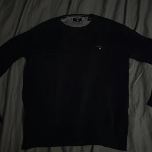 Svart stickad tröja från GANT - Svart stickad tröja från GANT med diskret logga på bröstet. Rund hals och ribbade muddar vid ärmslut och nederkant. Insidan av kragen har ett snyggt svartvitt mönster. Perfekt för dig som gillar klassisk och clean stil.