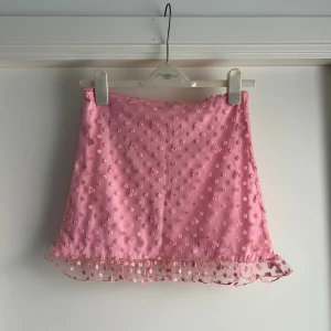 Rosa blommig minikjol från Sugar Thrillz - Supersöt rosa minikjol från Sugar Thrillz med små broderade blommor och volangkant i mesh. Kjolen har en lätt glansig yta och är perfekt för dig som gillar en gullig och lekfull stil. Sitter tajt och har en feminin vibe.