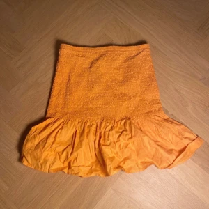 Orange volangkjol med smock - Superfin orange kortkjol med smock upptill och bred volang nertill. Kjolen har en härlig färg och är perfekt för dig som vill sticka ut. Den är lätt och luftig, vilket gör den skön att bära under varma dagar.