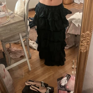 Svart långkjol med volanger - Säljer en svart långkjol med flera lager av stora volanger som ger ett riktigt flowy och coolt intryck. Kjolen har resår i midjan och är gjord i ett lätt, mjukt tyg som känns skönt mot huden. Perfekt för dig som gillar statement-plagg och vill sticka ut.
