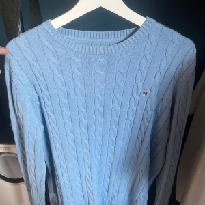 Ljusblå kabelstickad tröja från GANT - Säljer en ljusblå kabelstickad tröja från GANT i storlek L. Tröjan är tillverkad i mjuk bomull och har ett klassiskt kabelmönster samt rund halsringning. GANT-loggan med broderad sköld finns på bröstet. Perfekt för dig som gillar preppy stil.