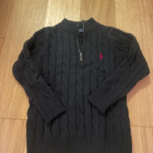 Svart 1/4 zip Polo Ralph Lauren - Svart kabelstickad tröja från Polo Ralph Lauren med halv dragkedja och röd broderad logga på bröstet. Tröjan har ribbade muddar och hög krage, perfekt för att hålla värmen. Klassisk och stilren design som funkar till många looks.
