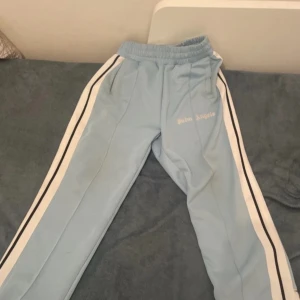 Palm Angels ljusblå mjukisbyxor - Snygga ljusblå mjukisbyxor från Palm Angels med vita och svarta ränder längs sidorna. Byxorna har elastisk midja och logga broderad på ena benet. Perfekta för en chill och sportig look.