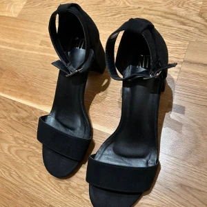 Svarta sandaler med klack och ankelrem - Fina svarta sandaler med bred rem över tårna och justerbar ankelrem med spänne. Materialet är av mocka. Använda ett fåtal gånger. 