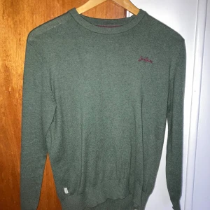 Grön stickad tröja från Scalpers - Mörkgrön stickad tröja från Scalpers med broderad logga framtill och en stor vinröd dödskalle på ryggen. Tröjan har rund halsringning och långa ärmar, perfekt för dig som gillar en stilren men edgy look.