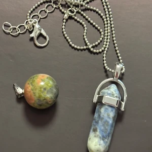 Halsband med stenar och silverkedja - Unikt halsband med två olika stenhängen – ett runt i grönt, orange och grått samt ett avlångt blåvitt kristallhänge med silverfäste. Kedjan är i silverfärgad metall med kulkedja och karbinlås. Perfekt för dig som gillar naturstenar och coola detaljer.