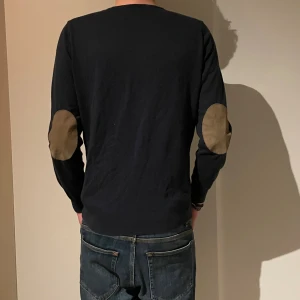 Massimo dutti tröja  - Svart långärmad tröja med rund hals och snygga beige mockalappar på armbågarna. Stilren och enkel design som passar perfekt till jeans. Materialet känns mjukt och skönt mot huden, och passformen är normal. Perfekt för dig som gillar klassiska detaljer.