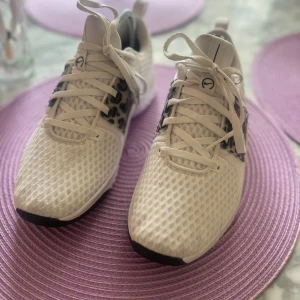 Nike Renew sneakers med leoparddetaljer - Snygga vita Nike Renew sneakers med mesh-ovandel och coola leopardmönstrade detaljer vid snörningen. Skorna har svart Nike-logga på sidan, platt sula och rund tå. Perfekta för dig som vill sticka ut med en sportig och trendig look.