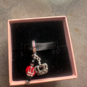 Charm till armband med krona och vakt - Unik charm till armband med två hängande detaljer: en brittisk vakt i röd och svart uniform samt en silverfärgad krona med små pärlor. Fästet har Union Jack-mönster i rött, blått och vitt. Perfekt för dig som gillar London vibes och detaljerade smycken.